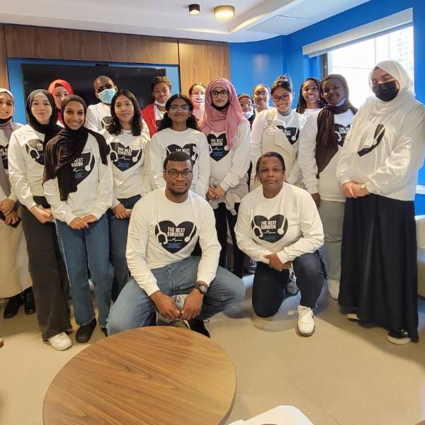 A group of high school students, program leaders and  Dr. Robert Yanagawa standing in a room wearing shirts with The Next Surgeon logo on the front.